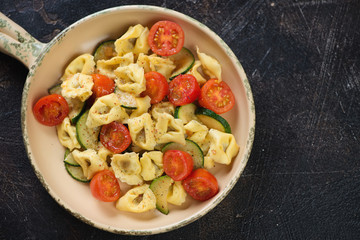 Top view of a serving pan with tortellini, roasted cherry tomatoes and zucchini. Horizontal shot on a dark brown stone background with space