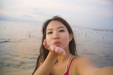 young happy and beautiful Asian Korean tourist woman taking self portrait selfie with mobile phone smiling having fun at sunset beach