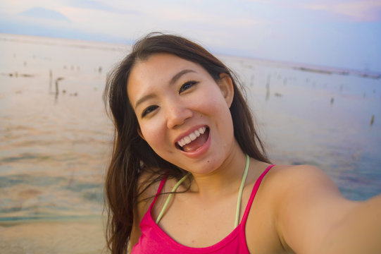 young happy and beautiful Asian Korean tourist woman taking self portrait selfie with mobile phone smiling having fun at sunset beach
