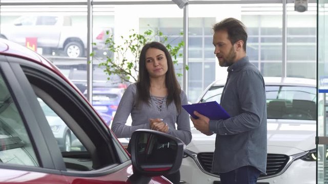 Mature bearded professional salesman working at the car dealership. Car dealer helping his female customer choosing a new automobile. Buying, communication, discount, advising.
