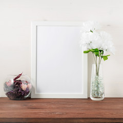 White frame, flower in glass bottle on dark brown wooden table against the white wall with copy space. Mock up.