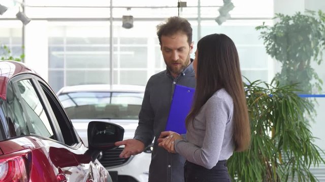 Professional salesman talking to a young woman at the car dealership salon. Female customer choosing a new automobile at the showroom. Mature auto dealer opening car door for a client.