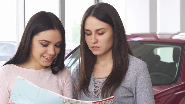 Close up of two beautiful happy women enjoying travelling together looking for destinations on a map standing in front of the car. Tourism, driving, navigation, lifestyle, vacation concept.