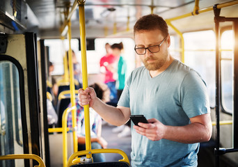 Young serious man is using his phone while standing in a bus and holding onto the yellow bar.