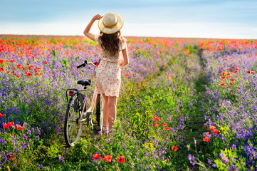 Romantic young woman standing by the back with bicycle on a poppy meadow