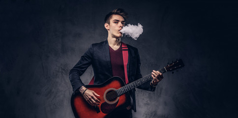 Handsome young musician with stylish hair in elegant clothes exhales smoke while playing acoustic guitar.