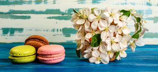 Multicolored macaroons and cup decorated with white flowers of apple trees on a blue wooden background.