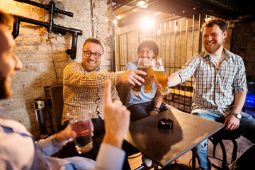 Cheerful male friends clinking with draft beer in front of their friend with a glass of water in...