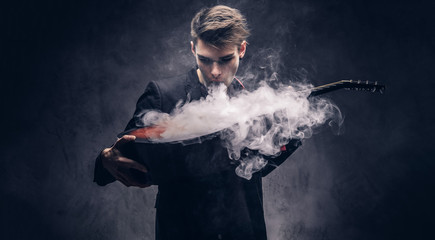 Handsome young musician with stylish hair in elegant clothes exhales smoke on his acoustic guitar.