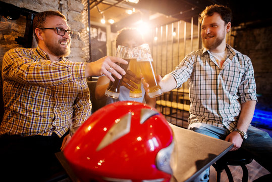Happy male friends clinking with draft beer while motorcycle helmet standing on a table in the local bar.