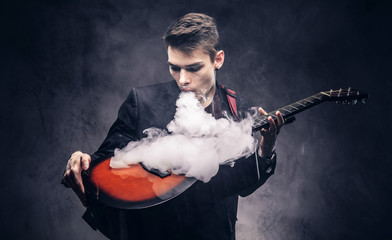 Handsome young musician with stylish hair in elegant clothes exhales smoke on his acoustic guitar.