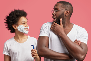 Surprised dark skinned African American male looks surprisingly at his teenager son who learns to...