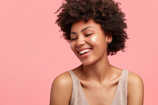 Sideways shot of cheerful slim lady with Afro hairstyle, has spangles on cheeks, smiles broadly, demonstrates white perfect teeth, dressed in casual t shirt, looks happily down, poses over pink wall