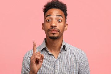 Studio shot of clever hipster with dark skin raises fore finger with intelligent look, gets...