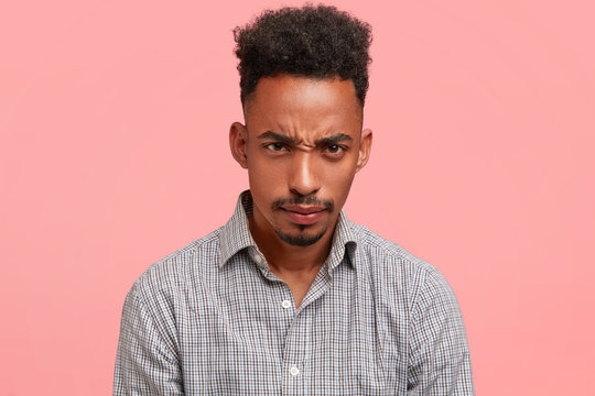 Negative facial expressions concept. Indoor shot of sullen grumpy African American male with trendy haircut, looks with displeased expression, being annoyed and unhappy, has much work on weekends