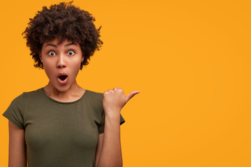 Photo of greatly surprised dark skinned female with bushy Afro haircut, wears casual t shirt, indicates at yellow blank copy space, keeps mouth widely opened, being astonished with something. - Powered by Adobe