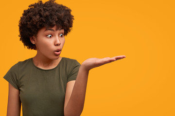 Horizontal shot of surprised African American woman indicates with palm away, shows something unexpected, stares with shock, isolated over bright yellow background. People, emotions, advertisement