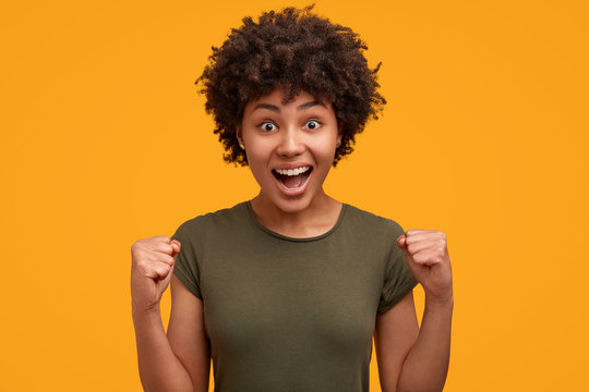 Bright image of overjoyed African American female with bushy hairstyle clenches fists like winner, exclaims with happiness, opens mouth widely, isolated over yellow background. Ethnicity concept