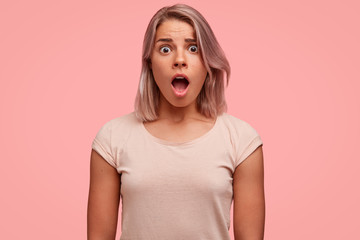 Image of frightened young European pretty female has wide opened mouth, stares at camera with bugged eyes, being scared by someone, expresses her big surprisement and astonishment, dressed casually.