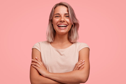 People, happiness and facial expressions concept. Pretty young woman with broad shining smile, keeps hands crossed, being in high spirit, wears braces on teeth, poses alone against pink background