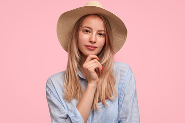 Elegant gorgeous lady wears stylish hat and shirt, looks with serious confident expression, listens something attentively, poses against pink background. Young female teacher being intelligent