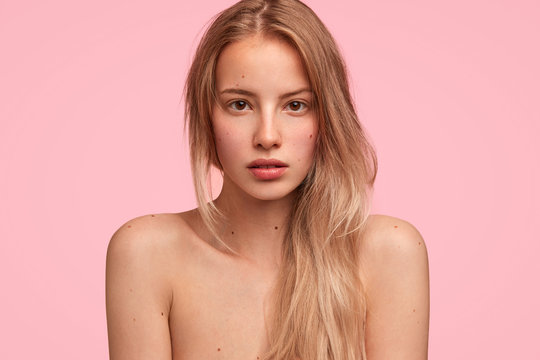 Close up shot of pleasant looking European female with confident expression, looks seriously directly at camera, poses nude against pink background, demonstrates her beauty and healthy skin.