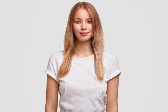 Portrait of pleased beautiful European female with long hair, dressed in casual t shirt, isolated on white background. Attractive woman student meets with groupmates. Facial expressions concept