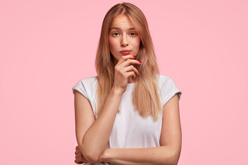 Confused thoughtful female with long hair, holds chin and looks in bewilderment, being puzzled as doesn`t understand something, isolated on pink studio background. Lovely youngster poses indoor - obrazy, fototapety, plakaty