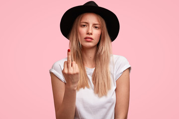 Fuck off! Studio shot of bad girl shows fuck sign, being dissatisfied with everything, isolated on pink background. Attractive woman in black hat and casual t shirt keeps middle finger raised