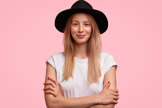 Elegant Caucasian lady wears stylish black hat and white t shirt, keeps arms folded, listens happily interlocutor, isolated on pink background. Delighted female tourist happy to explore new places