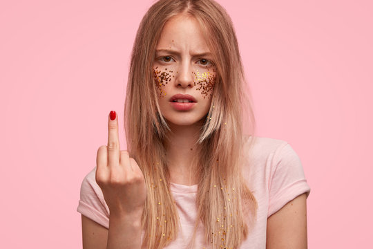 Annoyed female shows fuck sign, has argument with friend, looks with displeased expression at camera, isolated on pink studio background. Beautiful irritated young woman shows middle finger.