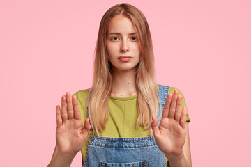 Young attractive female shows prohibition symbol or stop sign, refuses something, shows her refusal or rejection, dressed in jean overalls, poses against pink studio background. Body language concept