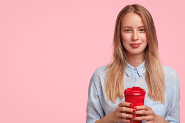 Lovely Caucasian female student has break after lectures, holds takeaway coffee, has pleasant talk with her groupmates, wears elegant shirt, poses against pink background with blank copy space