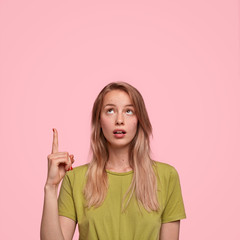 Indoor shot of good looking female with surprised expression, looks with bated breath at blank copy space, wears casual t shirt, isolated over pink background, copy space for your advertisement