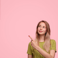 Vertical shot of good looking female model advertises new product in studio, points at blank copy space, isolated over pink background. Pretty woman seller attracts your attention to something
