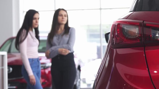 Selective focus on car lights on the foreground copy space. Two female customers discussing cars for sale at the dealership showroom. Consumerism, buying auto concept.