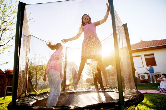 Two adorable playful girls and a little toddler boy are having time of their life on a trampoline while the rest of their family is grilling and having fun in a field.