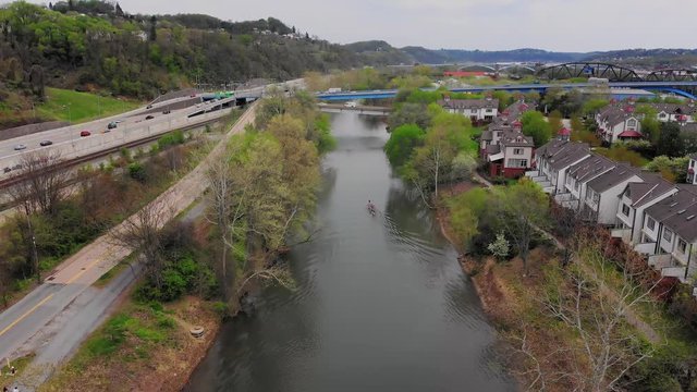 A slow reverse aerial establishing shot of kayakers on the Allegheny River on an overcast Spring day. Upscale homes seen on Washington's Landing.  	
