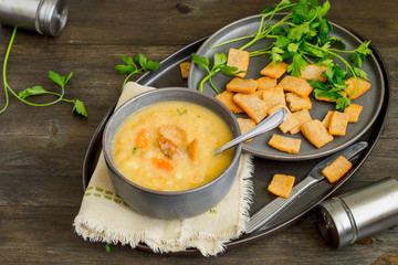 Pea thick soup in a bowl, next to fresh green parsley and small crispy croutons on a round dish. 