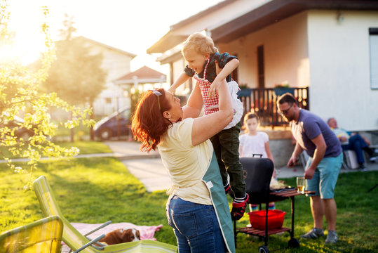 Woman is lifting up high her gorgeous and cheerful toddler boy while rest of the family is grilling.