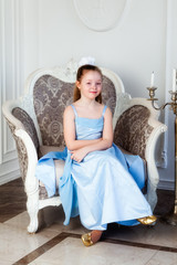 Girl in a blue dress is sitting in a armchair in a room and smiling
