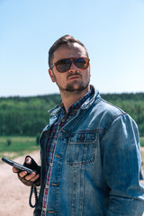 Portrait young guy with sunglasses and Jeans jacket uses a smartphone, holds in hands, vintage retro motorbike background, fashion men. old timer age concept, 1960s style, vertical photo