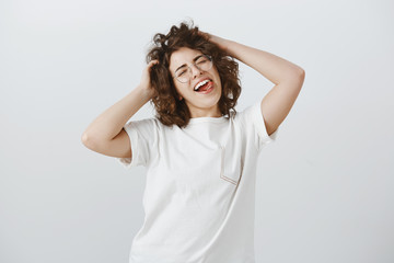 Enjoying being wild sometimes. Portrait of cheerful carefree young woman, making cute faces, tilting head and holding hands on hair, sticking out tongue playfully, having fun over gray background
