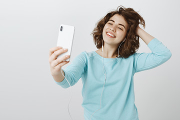 Fototapeta premium Time for selfie. Portrait of stylish confident good-looking female student in earphones, pulling hand with smartphone, taking photo and touching hair playfully, being happy over gray wall