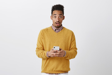 Someone tried to hack his phone. Shocked handsome boyfriend with afro hairstyle in trendy clothes holding smartphone, staring at camera with amazed expression, being surprised over gray wall