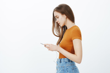 Fototapeta premium Profile shot of good-looking busy freelancer in stylish clothes, holding smartphone and listening music in earphones, sending message to boyfriend, having importrant conversation about relationship