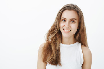 Positive charming caucasian female student with fair hair, tilting head and smiling friendly, listening carefully to interesting conversation, being happy and pleased while standing over gray wall