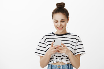 Girl editing photo to post it in social network. Focused pleased confident woman with bun hairstyle, wearing earphones and looking at screen of smartphone, watching video over white background