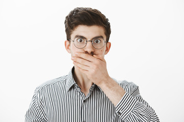 Classmate heard shocking news, being speechless. Portrait of amazed and stunned casual caucasian male model with eyebrows in trendy glasses, covering mouth with palm, being shocked and astonished