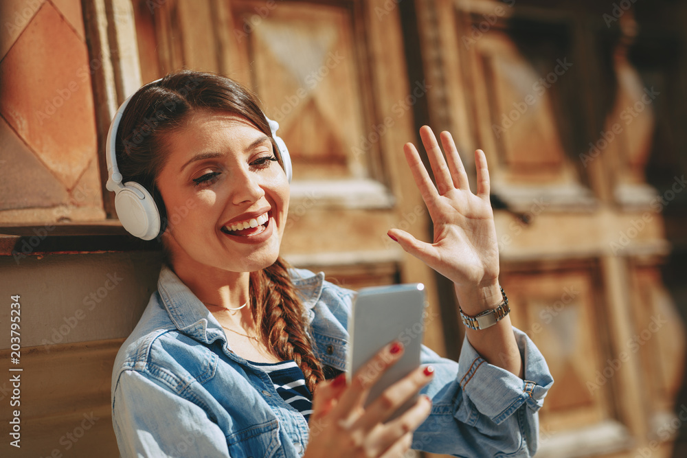 Wall mural Young woman making video call via smartphone and headphones  in the city